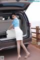 A woman in a white skirt and blue shirt is opening the trunk of a car.